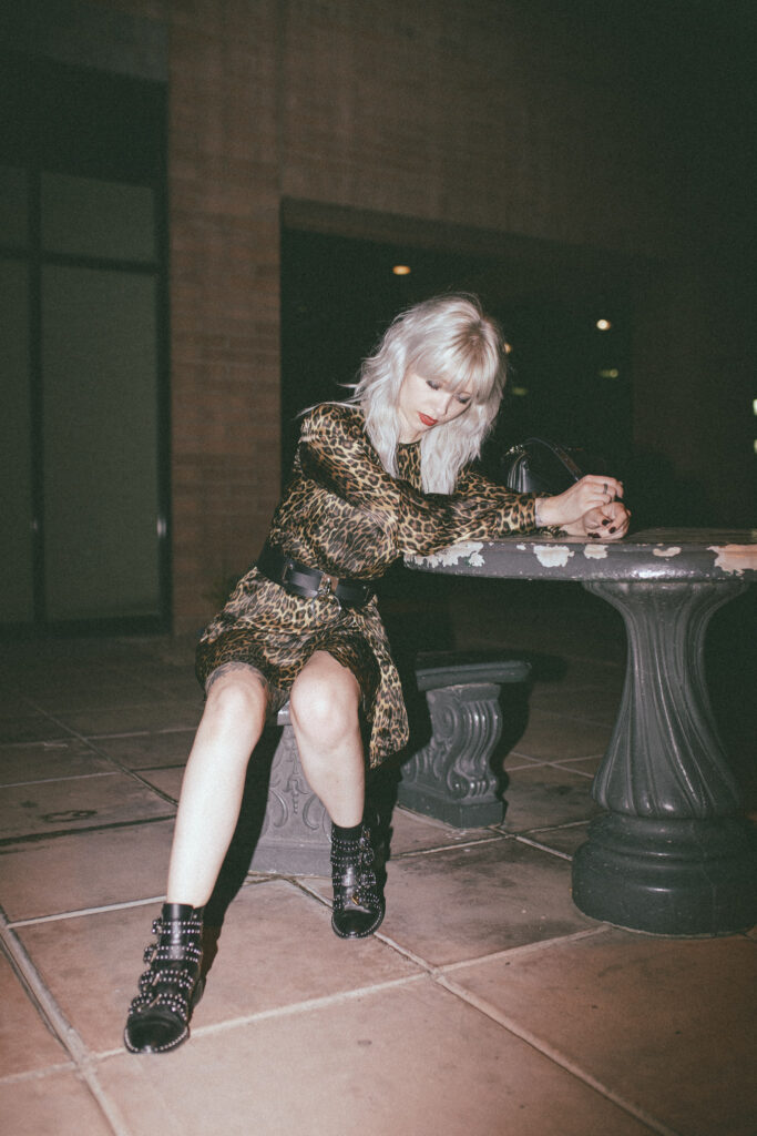 Trina in a leopard dress and studded boots sitting at a table and looking away from the camera