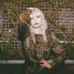 Trina in a leopard dress standing in bront of a brick wall with vines on it