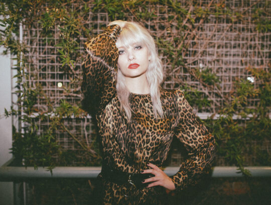 Trina in a leopard dress standing in bront of a brick wall with vines on it