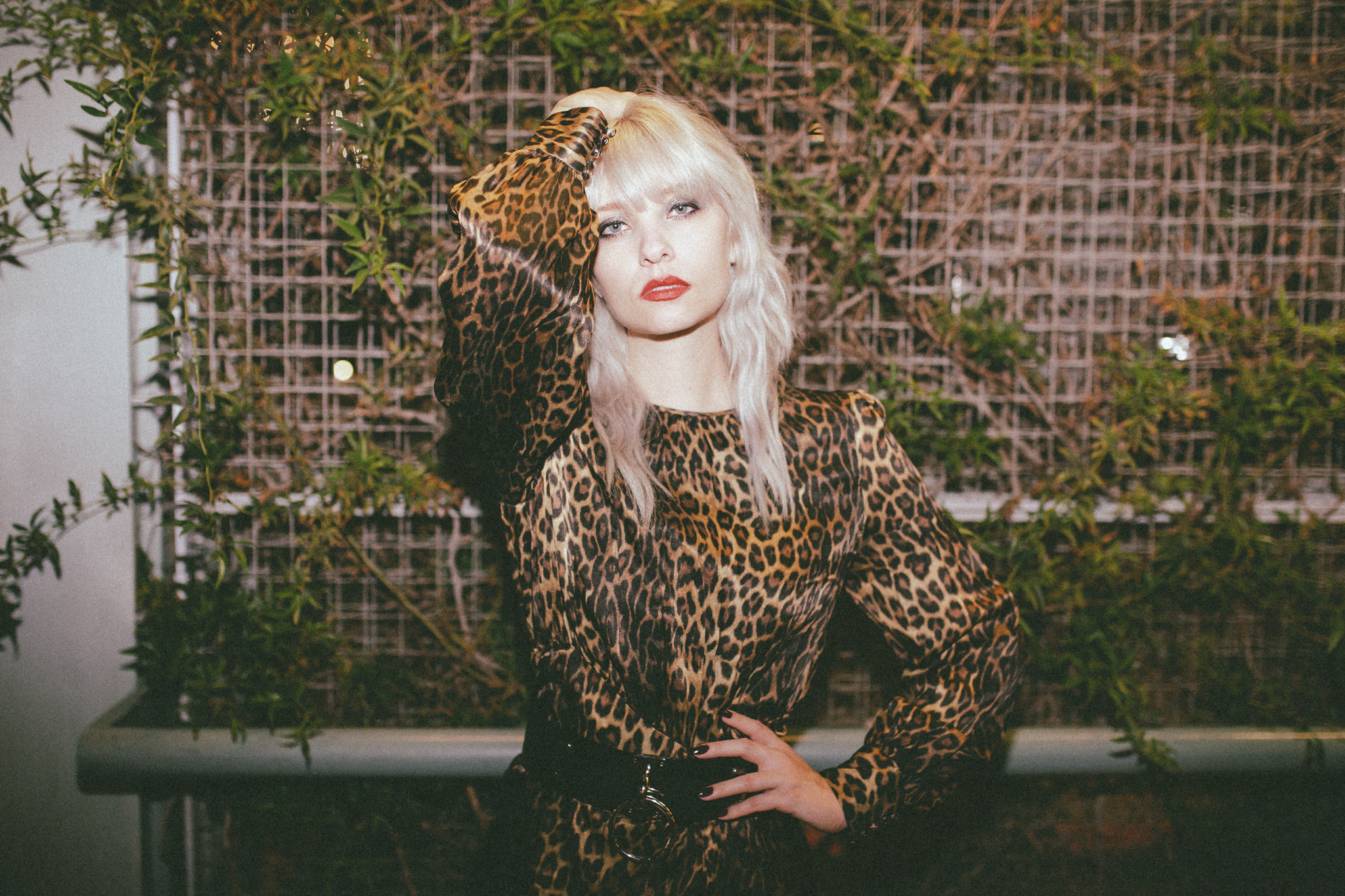 Trina in a leopard dress standing in bront of a brick wall with vines on it